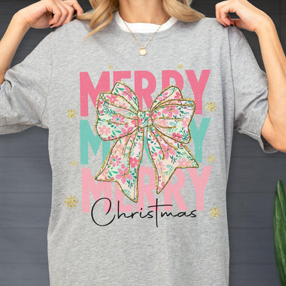 May include: A gray t-shirt with the words "MERRY CHRISTMAS" in pink and teal. A floral bow graphic is centered on the shirt. Gold glitter accents the design. The shirt has a white collar.