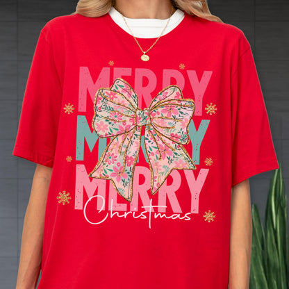 May include: Red t-shirt with a large floral bow graphic and the words "MERRY CHRISTMAS" in pink and white. The bow has pink and blue flowers with gold accents. Gold snowflakes are scattered around the design.