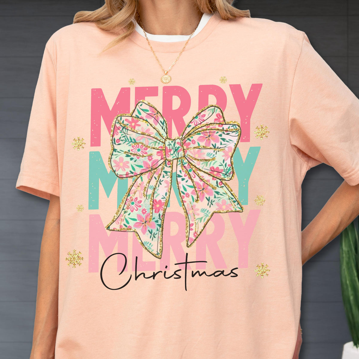 May include: A peach-colored t-shirt with a floral bow graphic that says "Merry Christmas." The bow is pink, green, and gold. The text "Merry" is in pink and the text "Christmas" is in black script.