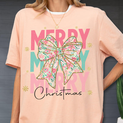 May include: A peach-colored t-shirt with a floral bow graphic that says "Merry Christmas." The bow is pink, green, and gold. The text "Merry" is in pink and the text "Christmas" is in black script.