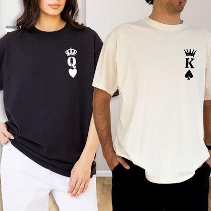 May include: A black t-shirt featuring a white queen of hearts playing card symbol on the chest and a cream t-shirt featuring a black king of spades playing card symbol on the chest. Both shirts have a crew neckline and short sleeves.