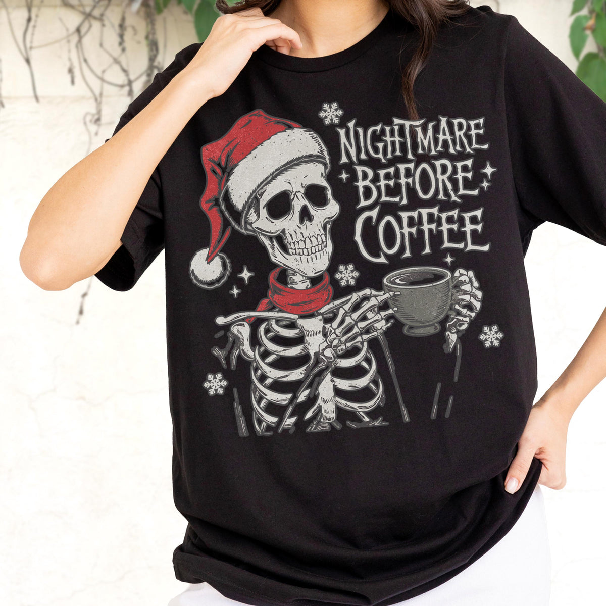 May include: Black t-shirt featuring a skeleton wearing a Santa hat and red scarf, holding a coffee cup. The text "NIGHTMARE BEFORE COFFEE" is printed above the skeleton. Snowflake graphics are also included in the design.