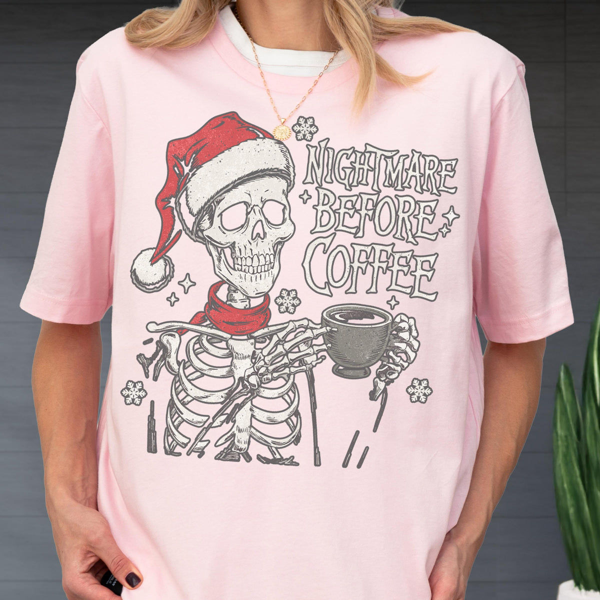 May include: A light pink t-shirt featuring a skeleton wearing a Santa hat and a red scarf, holding a coffee cup. The text "NIGHTMARE BEFORE COFFEE" is printed above the skeleton in a stylized font. Snowflakes are also included in the design.