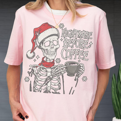 May include: A light pink t-shirt featuring a skeleton wearing a Santa hat and a red scarf, holding a coffee cup. The text "NIGHTMARE BEFORE COFFEE" is printed above the skeleton in a stylized font. Snowflakes are also included in the design.