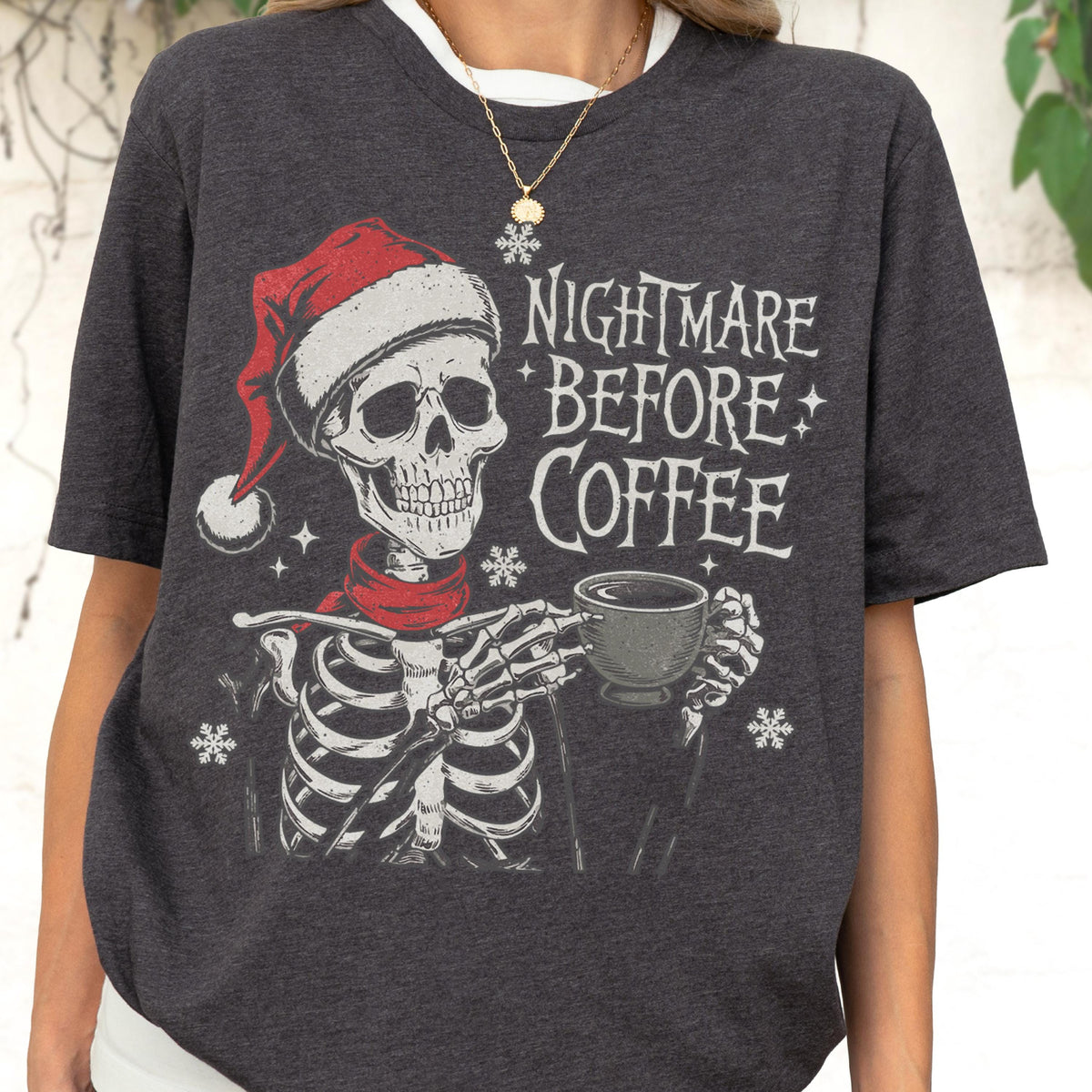 May include: Dark gray t-shirt featuring a skeleton wearing a Santa hat and red scarf, holding a coffee cup. The text "NIGHTMARE BEFORE COFFEE" is printed above the skeleton. Snowflake and star graphics are also included in the design.