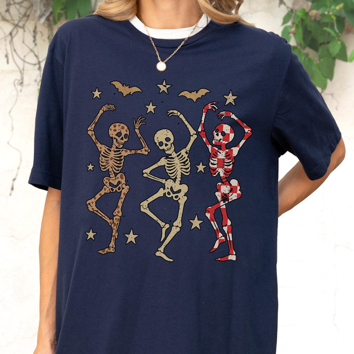May include: Navy blue t-shirt featuring a Halloween design. The graphic shows three dancing skeletons in various colors, surrounded by stars and bats. The shirt has a white crew neck and is a casual, comfortable style.