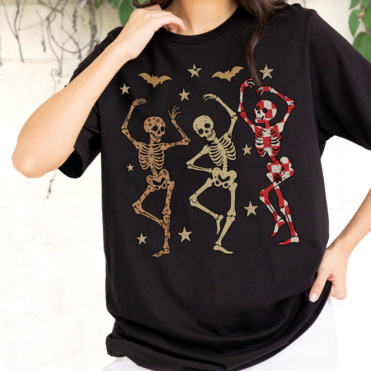 May include: Black t-shirt featuring a graphic of three dancing skeletons in various poses. The skeletons are in shades of brown and red, with stars and bat silhouettes scattered around them. A fun and festive design for Halloween.