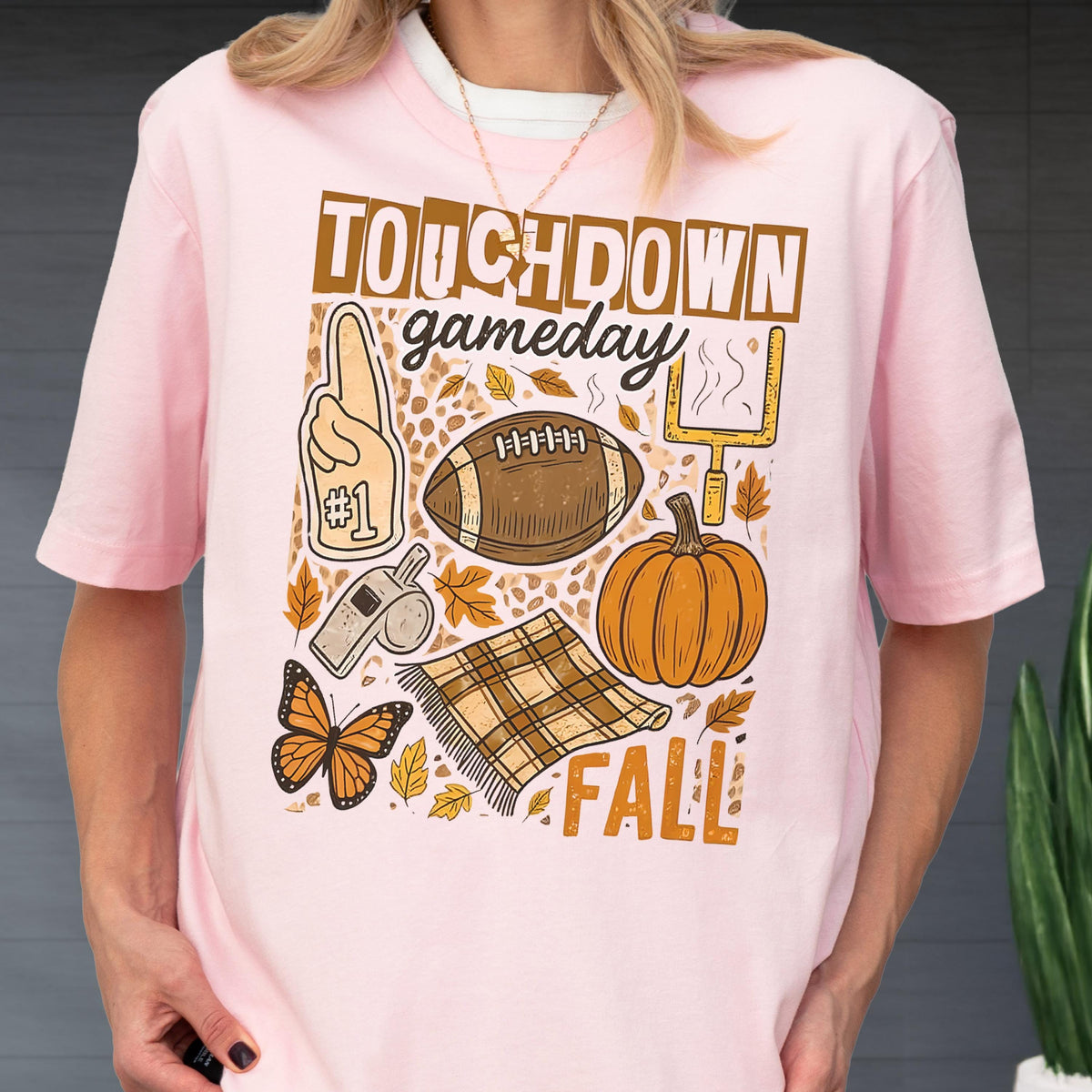 May include: Pink t-shirt with a fall-themed graphic. The design includes a football, pumpkin, and the words "TOUCHDOWN gameday FALL". Other elements include a foam finger, a whistle, a butterfly, and a plaid blanket.