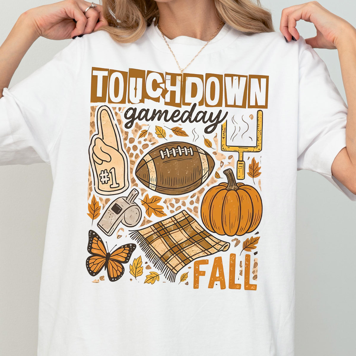 May include: White t-shirt with a fall-themed graphic. The design includes the words "TOUCHDOWN gameday FALL", a football, a pumpkin, a #1 foam finger, a whistle, a butterfly, and a plaid blanket.