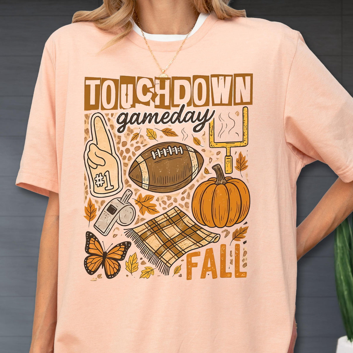 May include: A light peach-colored t-shirt with a fall-themed graphic. The design includes the words "TOUCHDOWN gameday FALL", a football, a pumpkin, a #1 foam finger, a whistle, a butterfly, and a plaid blanket.