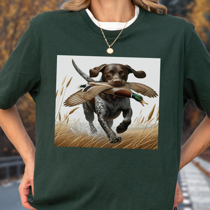 May include: Dark green t-shirt featuring a graphic of a dog running through tall grass with a duck in its mouth. The dog is brown and white, and the duck has brown, green, and white feathers. The background is a light gray.