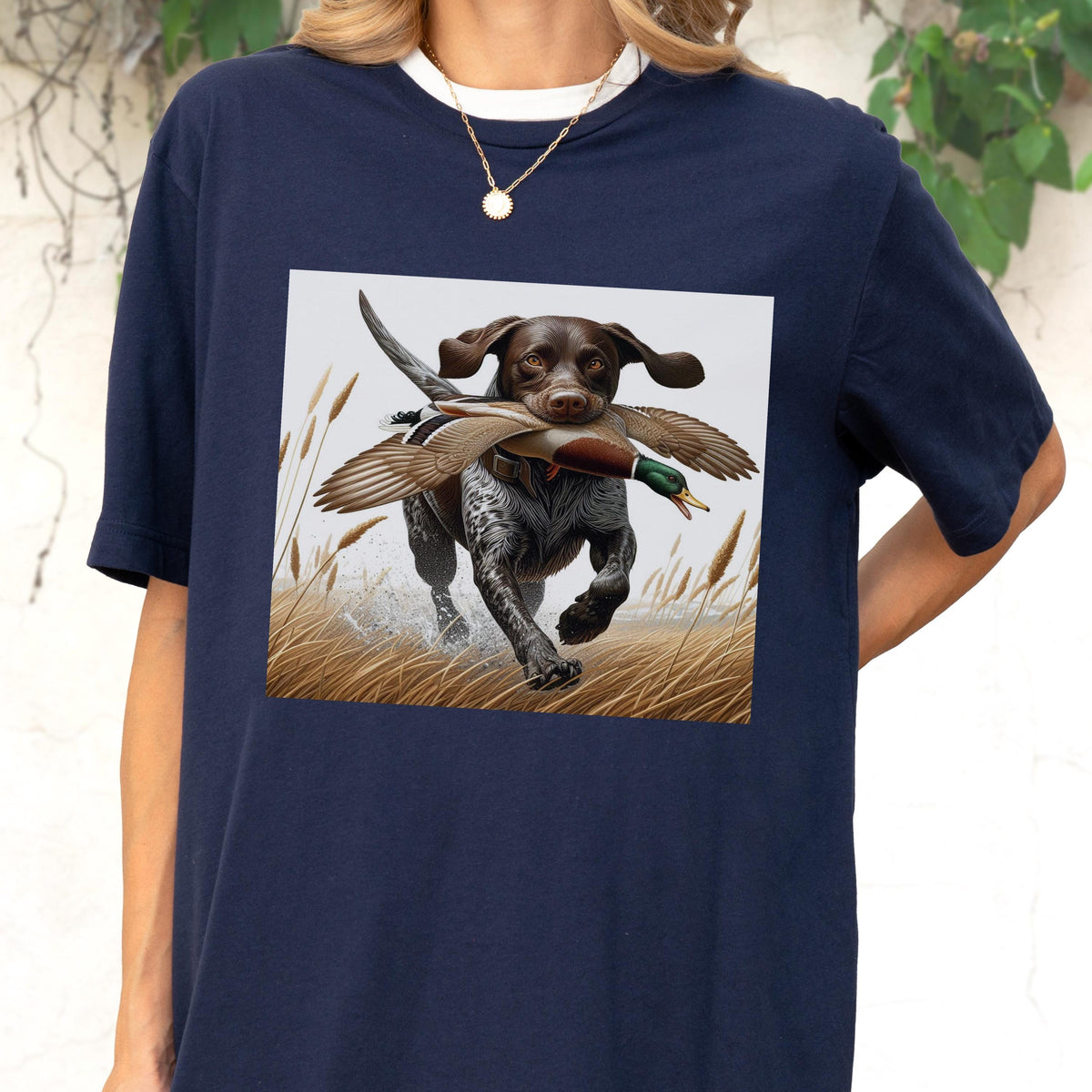 May include: Navy blue t-shirt featuring a square graphic of a brown dog with black spots running through tall grass, carrying a duck in its mouth. The dog has floppy ears and is running through water. The shirt has a white collar.