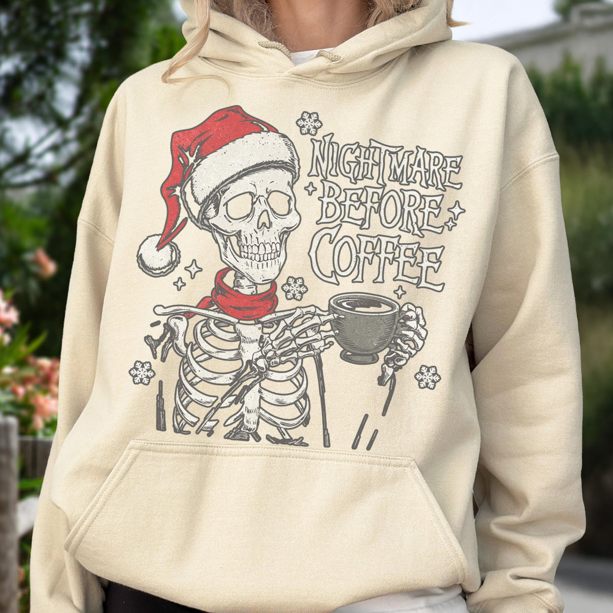 May include: Beige hoodie featuring a skeleton wearing a Santa hat and red scarf, holding a coffee cup. The text "NIGHTMARE BEFORE COFFEE" is printed above the skeleton. The design includes snowflake and star accents.