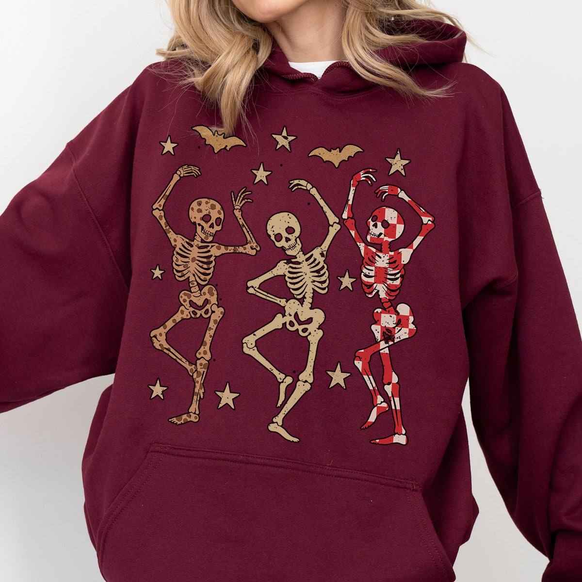 May include: Burgundy hoodie featuring a graphic of three dancing skeletons, bats, and stars. The skeletons are in various poses, with one skeleton in a red and white checkered pattern. The design is a fun, festive theme.