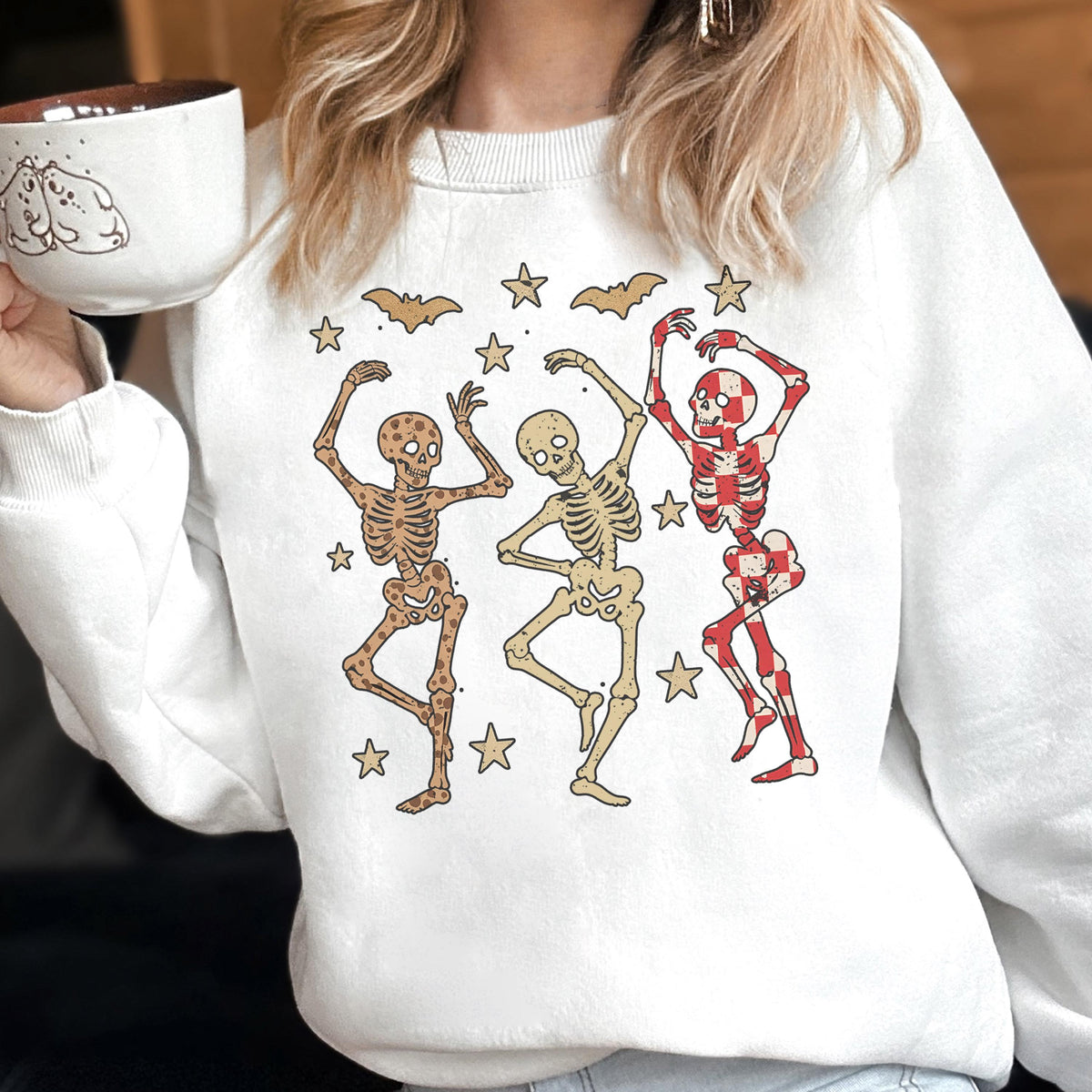 May include: White sweatshirt featuring a graphic of three dancing skeletons in brown, beige, and red. The design includes bats and stars, creating a festive Halloween theme. The sweatshirt is a comfortable, casual style.