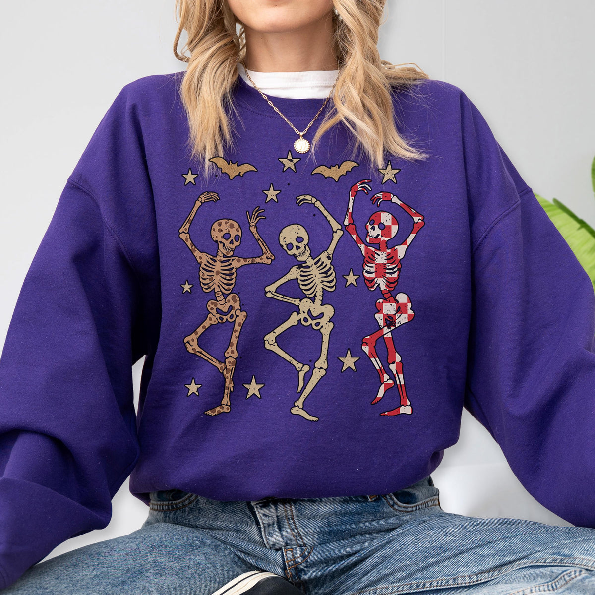 May include: A purple sweatshirt featuring a graphic of three dancing skeletons. The skeletons are in various poses, with one in a red and white checkered pattern. Gold stars and bats are scattered around the skeletons.