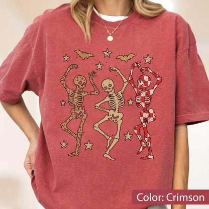 May include: Crimson-colored t-shirt featuring a graphic of three dancing skeletons, stars, and bats. The skeletons are in various poses, with one skeleton in a red and white checkered pattern. The text "Color: Crimson" is at the bottom.