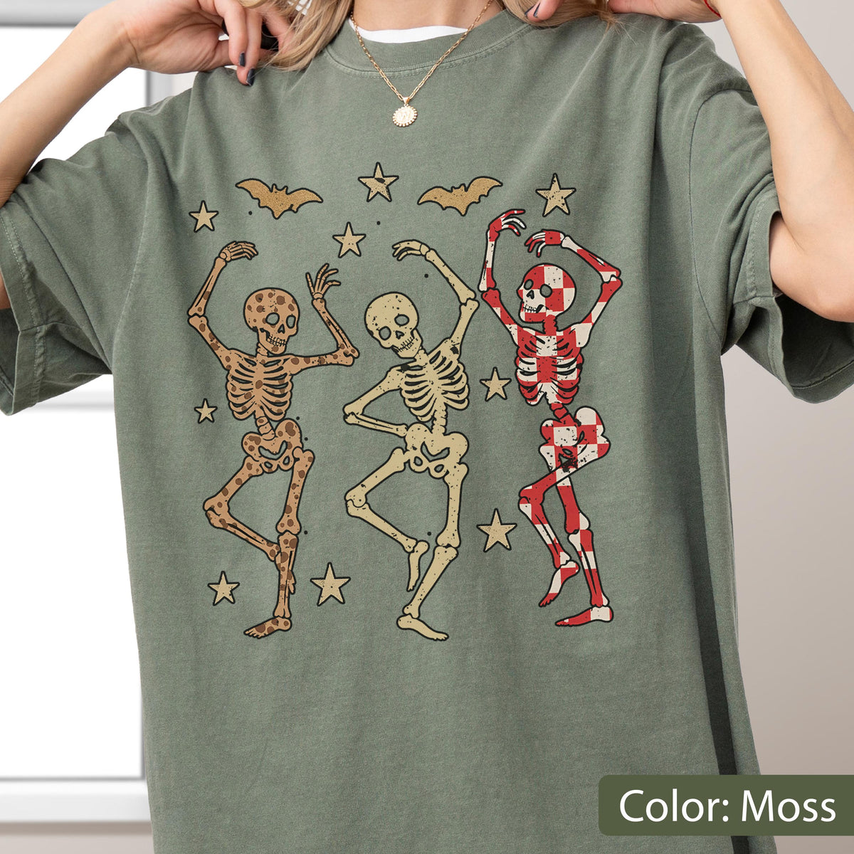 May include: Moss green t-shirt featuring a graphic of three dancing skeletons, bats, and stars. One skeleton is red and white checkered, another is tan with brown spots, and the third is tan. The text "Color: Moss" is at the bottom.