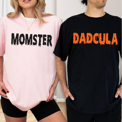 May include: A light pink t-shirt with the black text MOMSTER in a dripping font and a black t-shirt with the orange text DADCULA in a dripping font. Both shirts feature short sleeves and a crew neckline, displaying a graphic design style.