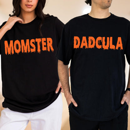 May include: Two black short-sleeve t-shirts. The left shirt features the orange text MOMSTER in a dripping font. The right shirt features the orange text DADCULA in a dripping font. Both shirts have a crew neckline and a standard fit.