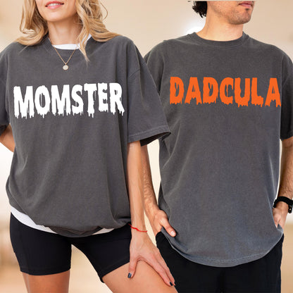 May include: Two charcoal gray short-sleeve t-shirts. The left shirt features the white text MOMSTER in a dripping font. The right shirt features the orange text DADCULA in a dripping font. Both shirts have a crew neckline and a relaxed fit.
