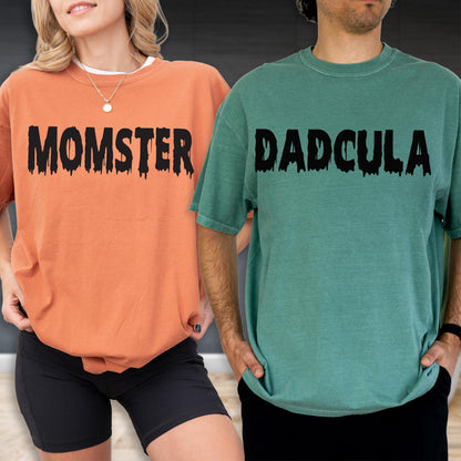 May include: An orange t-shirt with the black text MOMSTER in a dripping font and a green t-shirt with the black text DADCULA in a dripping font. Both shirts feature short sleeves and a crew neckline, showcasing a casual and relaxed fit.