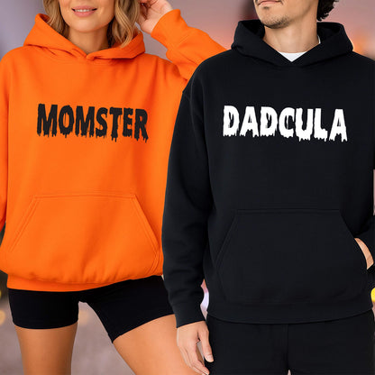 May include: Two hooded sweatshirts, one orange and one black. The orange sweatshirt has the word "MOMSTER" printed in black, and the black sweatshirt has the word "DADcula" printed in white. Both sweatshirts have a front pocket and a hood.