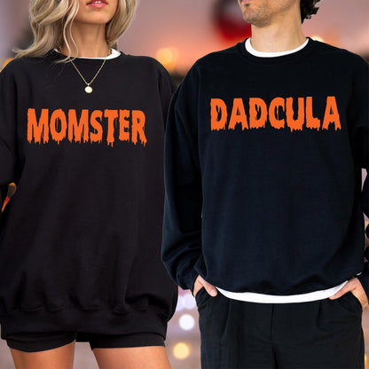 May include: Two black sweatshirts with orange text. One says "MOMSTER" and the other says "DADCULA". Both sweatshirts have a white crew neck collar. The sweatshirts are likely for a Halloween or spooky-themed event.