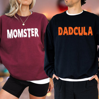 May include: Two sweatshirts, one burgundy and one navy blue. The burgundy sweatshirt has the word "MOMSTER" in white dripping font. The navy blue sweatshirt has the word "DADCULA" in orange dripping font. Both sweatshirts have a white crew neck.