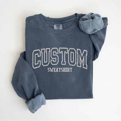 May include: A navy blue sweatshirt with the word "CUSTOM" in white, block-style lettering. Below the word "CUSTOM" is the word "SWEATSHIRT" in a smaller font. The sleeves are rolled up, revealing a lighter blue interior.