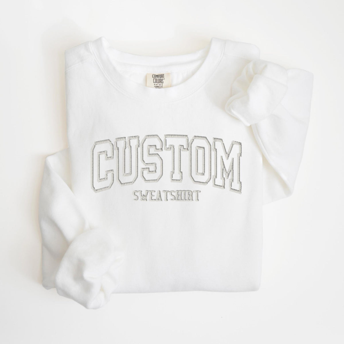 May include: A white sweatshirt with the word "CUSTOM" in a gray, outlined, collegiate-style font. Below the word "CUSTOM" is the word "SWEATSHIRT" in a smaller, gray font. The sweatshirt has a crew neck and long sleeves.