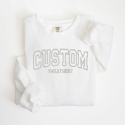 May include: A white sweatshirt with the word "CUSTOM" in a gray, outlined, collegiate-style font. Below the word "CUSTOM" is the word "SWEATSHIRT" in a smaller, gray font. The sweatshirt has a crew neck and long sleeves.