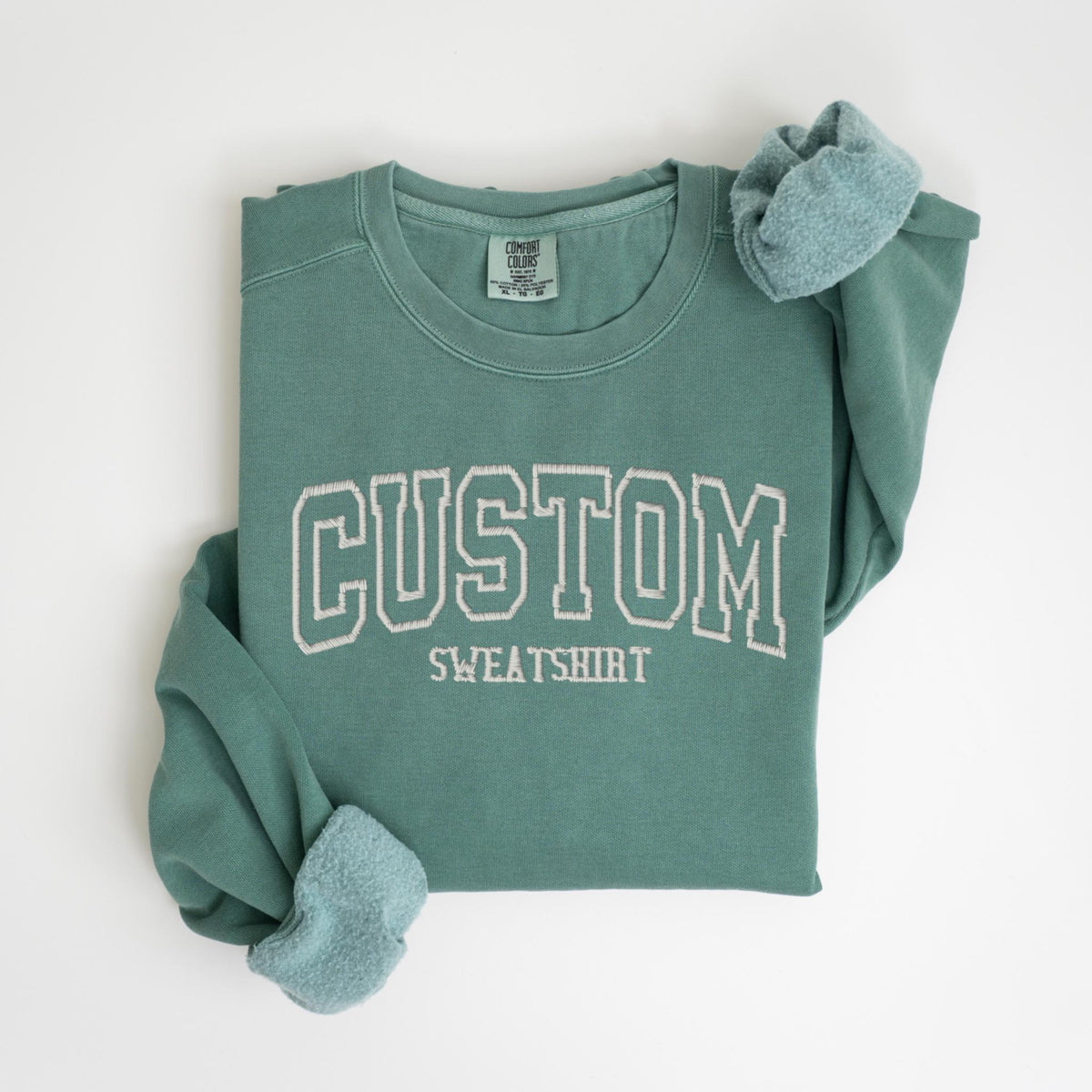 May include: A teal green sweatshirt with the word "CUSTOM SWEATSHIRT" embroidered in white. The sweatshirt is folded, with the sleeves showing a soft, light blue lining. The Comfort Colors tag is visible.