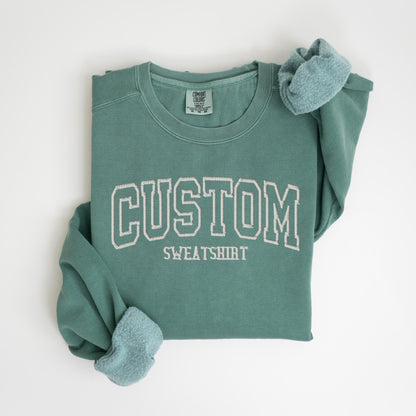May include: A teal green sweatshirt with the word "CUSTOM SWEATSHIRT" embroidered in white. The sweatshirt is folded, with the sleeves showing a soft, light blue lining. The Comfort Colors tag is visible.