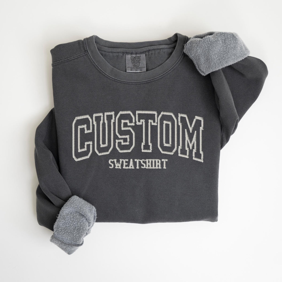 May include: Dark gray sweatshirt with the word "CUSTOM" in white, outlined lettering. The word "SWEATSHIRT" is below the word "CUSTOM". The sleeves are cuffed, revealing a soft, gray lining. The sweatshirt is a comfortable, casual style.