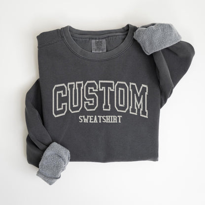 May include: Dark gray sweatshirt with the word "CUSTOM" in white, outlined lettering. The word "SWEATSHIRT" is below the word "CUSTOM". The sleeves are cuffed, revealing a soft, gray lining. The sweatshirt is a comfortable, casual style.
