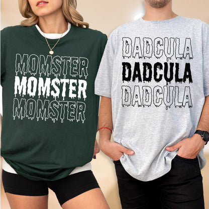 May include: Two t-shirts, one dark green and one gray, with Halloween-themed text. The dark green shirt says "MOMSTER" in white, dripping font. The gray shirt says "DADcula" in a similar font. Both shirts have a white collar.