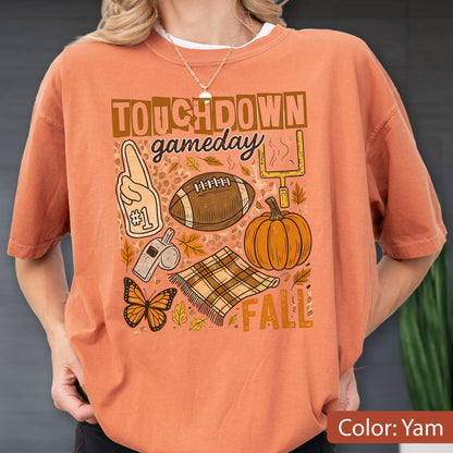 May include: Orange-colored t-shirt with a graphic design featuring the words "TOUCHDOWN gameday FALL". The design includes a football, pumpkin, and other fall-themed elements. The shirt is a solid color.