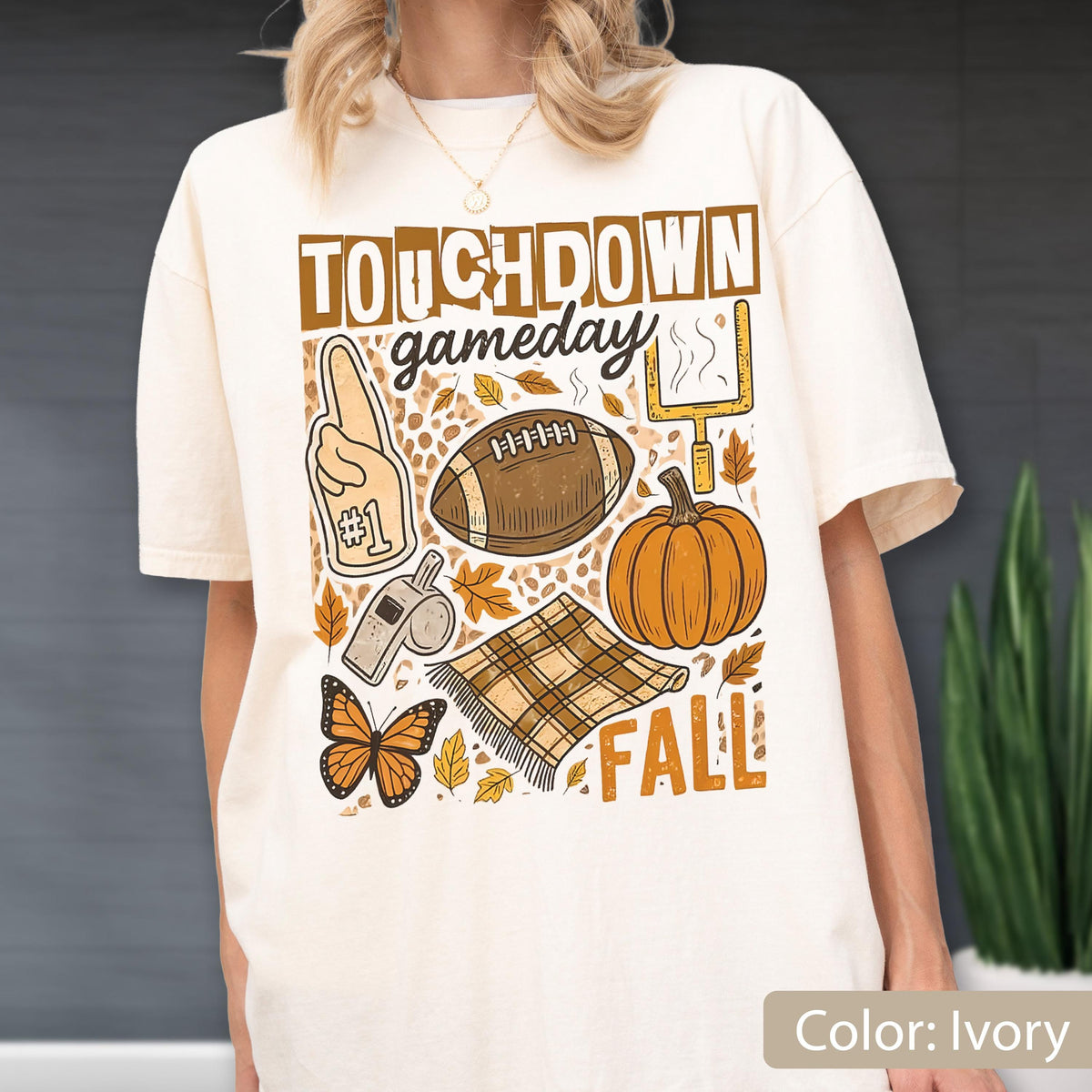 May include: Ivory-colored t-shirt with a fall-themed graphic. The design includes the words "TOUCHDOWN gameday FALL", a football, a pumpkin, a foam finger, a whistle, a butterfly, and autumn leaves. The shirt is a relaxed fit.