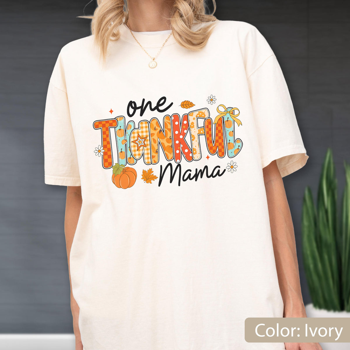 May include: Ivory-colored t-shirt with the text "one THANKFUL Mama" in a colorful, fall-themed design. The design includes pumpkins, leaves, and floral accents. The shirt is a relaxed fit.