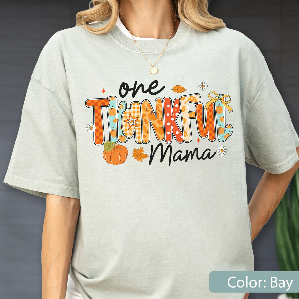 May include: A light green t-shirt with the words "one THANKFUL Mama" in colorful, patterned letters. The design includes pumpkins, leaves, and flowers, perfect for the fall season. The shirt is a relaxed fit.