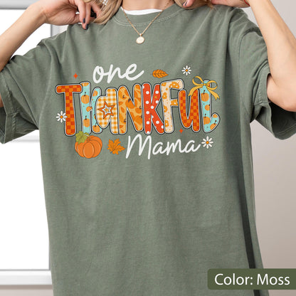 May include: Moss green t-shirt with the text "one THANKFUL Mama" in a colorful fall-themed design. The word "THANKFUL" features orange and blue patterns with pumpkin and leaf accents. A pumpkin and flowers are also included.
