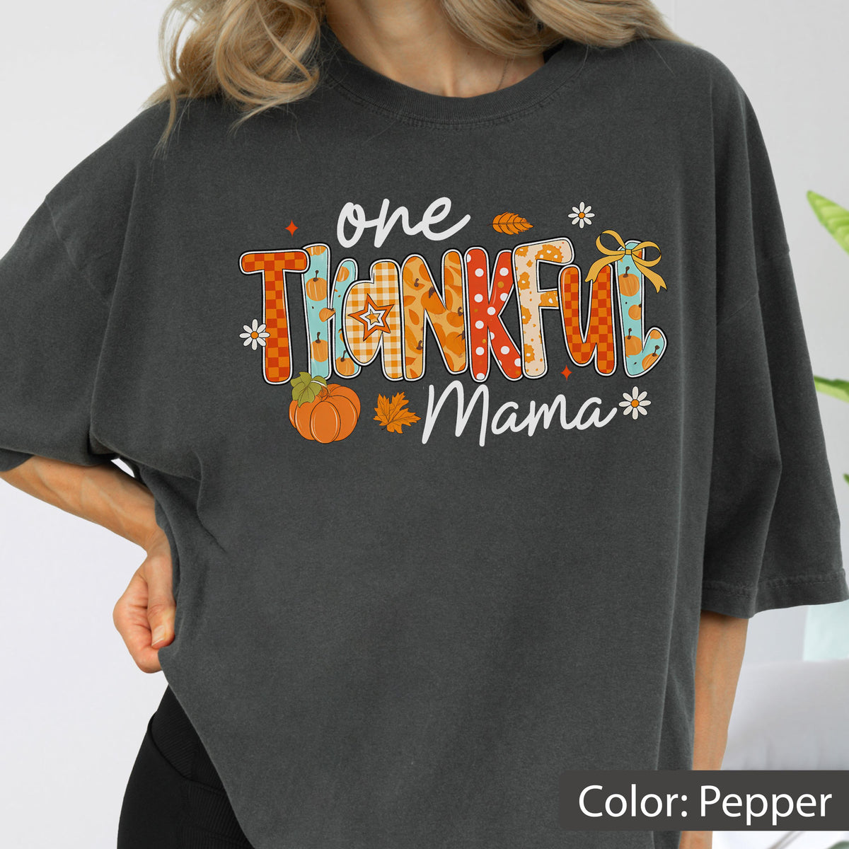 May include: A dark gray t-shirt with the words "one THANKFUL Mama" in a colorful, fall-themed design. The design includes pumpkins, leaves, and floral accents, perfect for Thanksgiving or autumn.