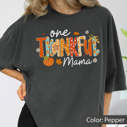 May include: A dark gray t-shirt with the words "one THANKFUL Mama" in a colorful, fall-themed design. The design includes pumpkins, leaves, and floral accents, perfect for Thanksgiving or autumn.