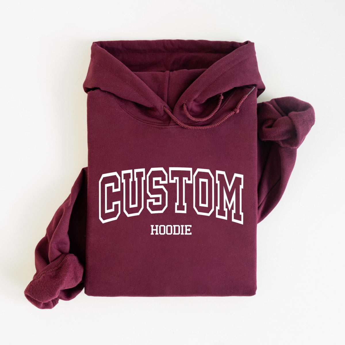May include: A maroon hoodie with the word "CUSTOM" in white, outlined block letters. Below the word "CUSTOM" is the word "HOODIE" in a smaller, white font. The hoodie is folded, with the hood up and the sleeves folded.