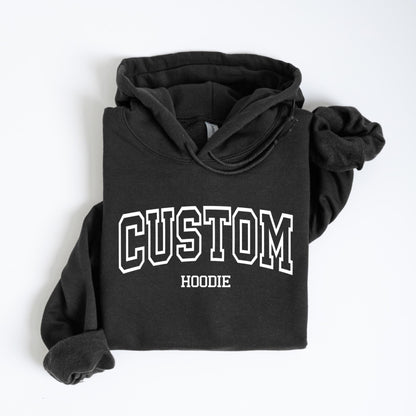 May include: A dark gray hoodie with the word "CUSTOM" in large white block letters across the chest, with "HOODIE" below. The hoodie has a drawstring hood and folded sleeves.