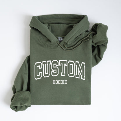 May include: An olive green hoodie with white text that reads "CUSTOM HOODIE" in a collegiate font. The hoodie has a drawstring hood and long sleeves. The garment is folded neatly on a white surface.
