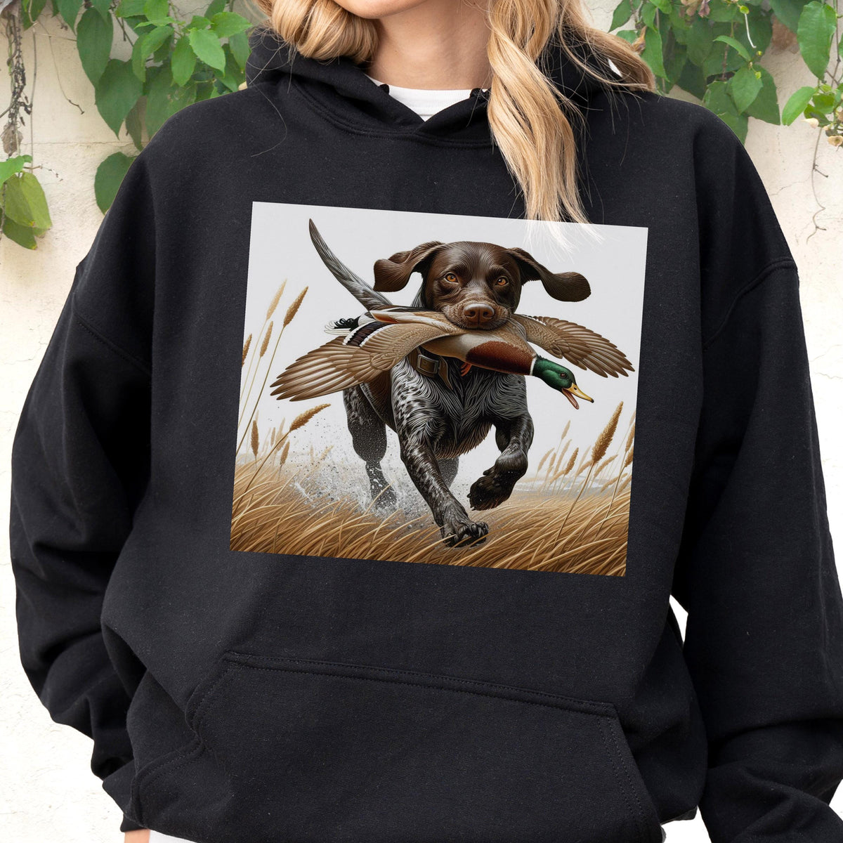 May include: Black hoodie featuring a graphic of a brown and white dog running through tall grass, carrying a duck in its mouth. The dog is depicted in a dynamic pose, with water splashing around its paws. The design is set against a neutral background.