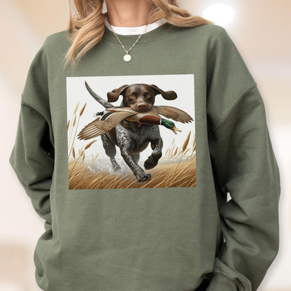 May include: Green sweatshirt featuring a graphic of a brown and white dog running through tall grass, carrying a duck in its mouth. The duck has brown and green feathers. The dog is the focal point of the design.