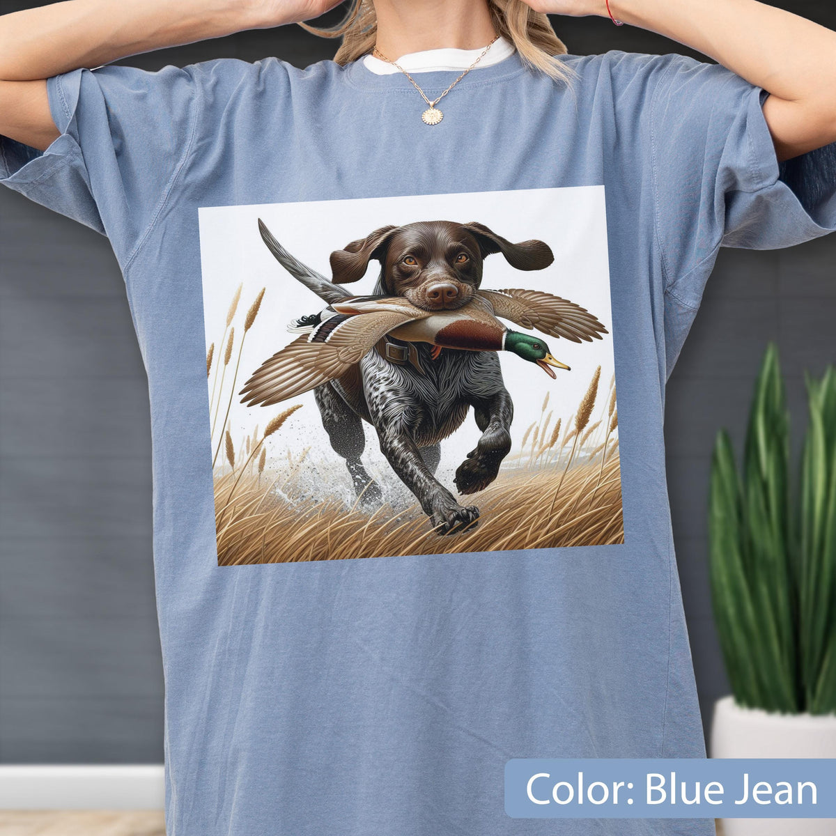 May include: A blue jean colored t-shirt featuring a graphic print of a brown and white spotted hunting dog running through tall golden grass while carrying a mallard duck in its mouth. The dog has floppy ears and a focused expression.